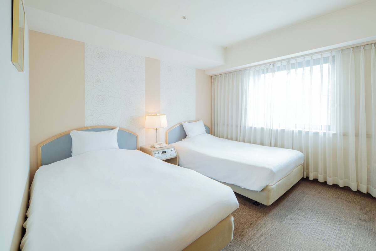 Een hotelkamer met twee bedden, een nachtkastje, een lamp en een groot raam met gordijnen.