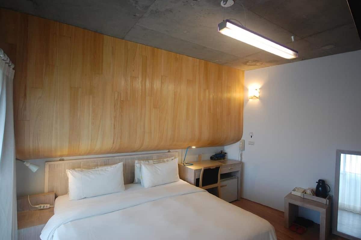 A hotel room with a wooden headboard, a bed with white linens, a desk with a lamp, a small table with a phone and a kettle, and a window with curtains.