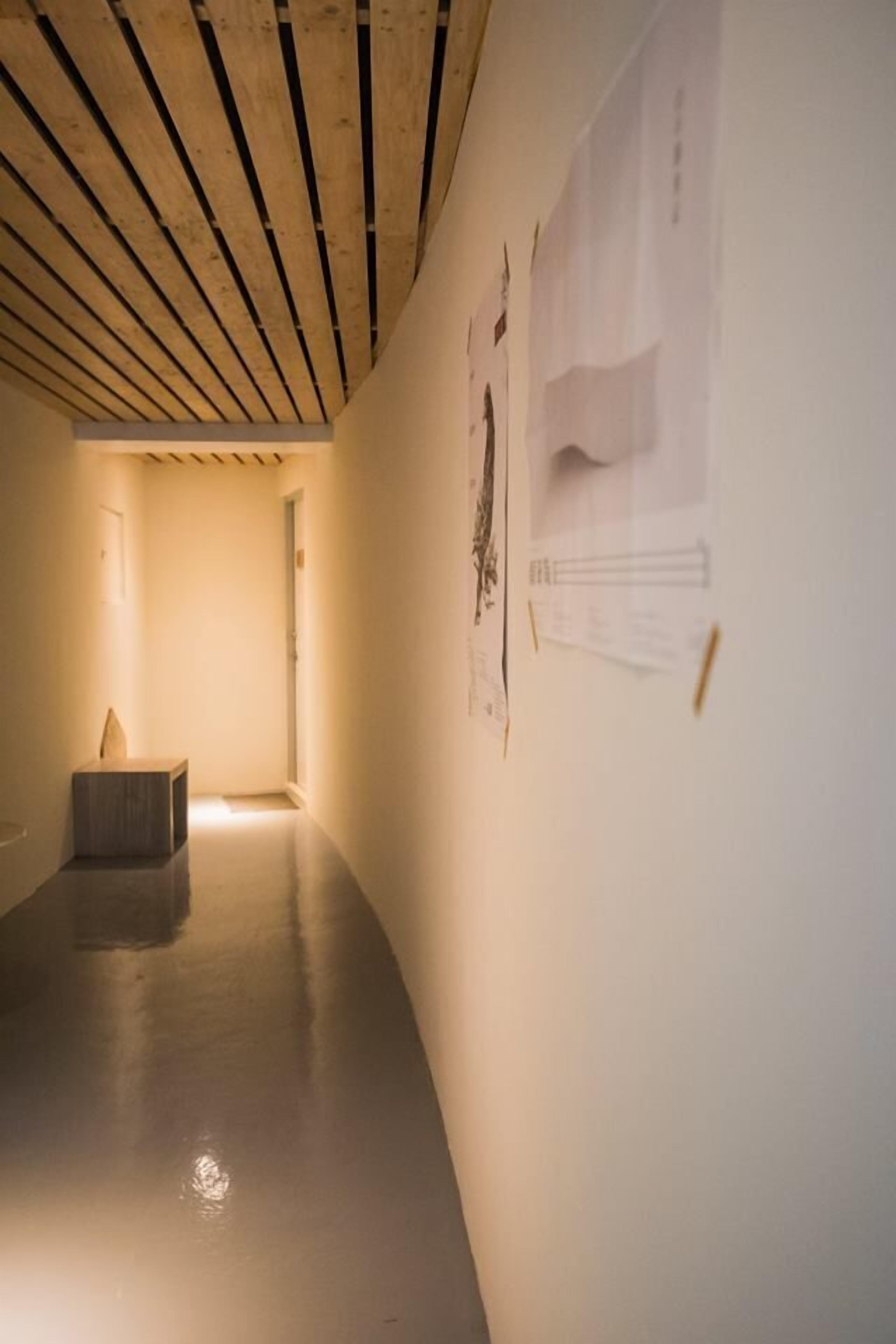 A hallway with a wooden ceiling, a small seating area, and walls with mounted papers.
