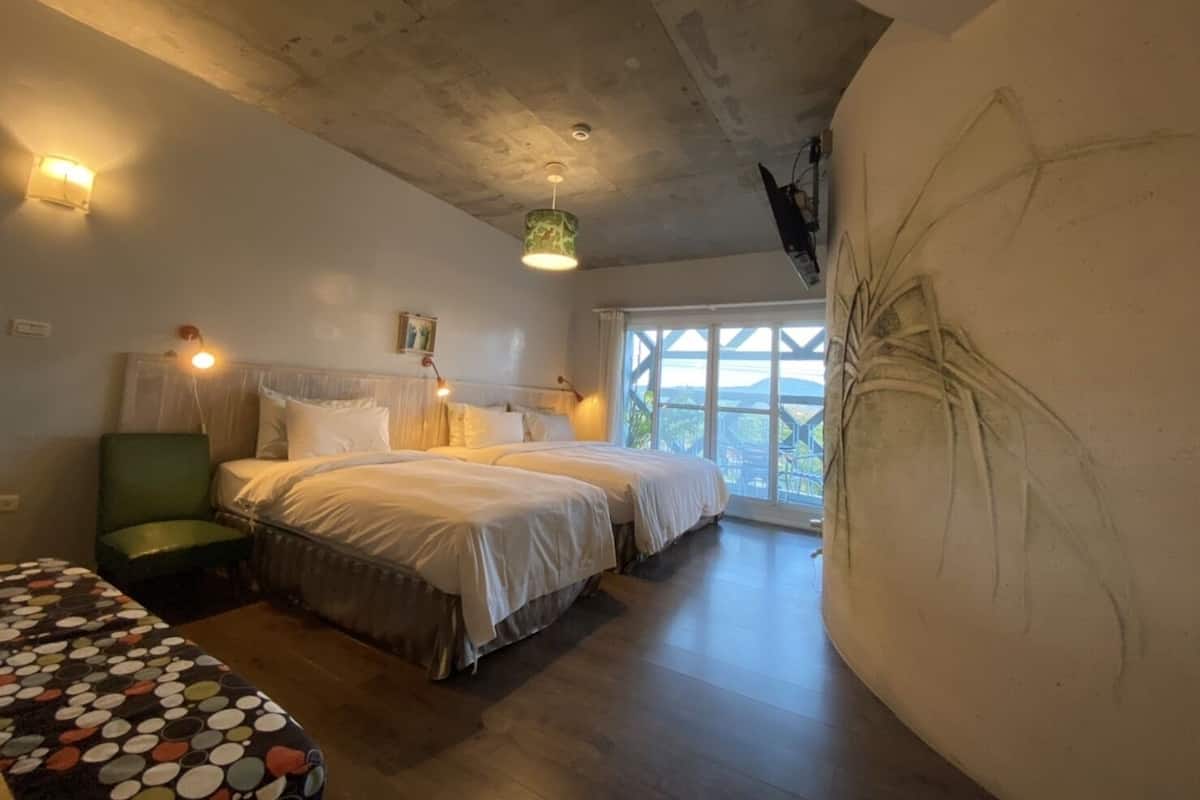 A bedroom with two beds, a green chair, a patterned table, and a large window with a view.