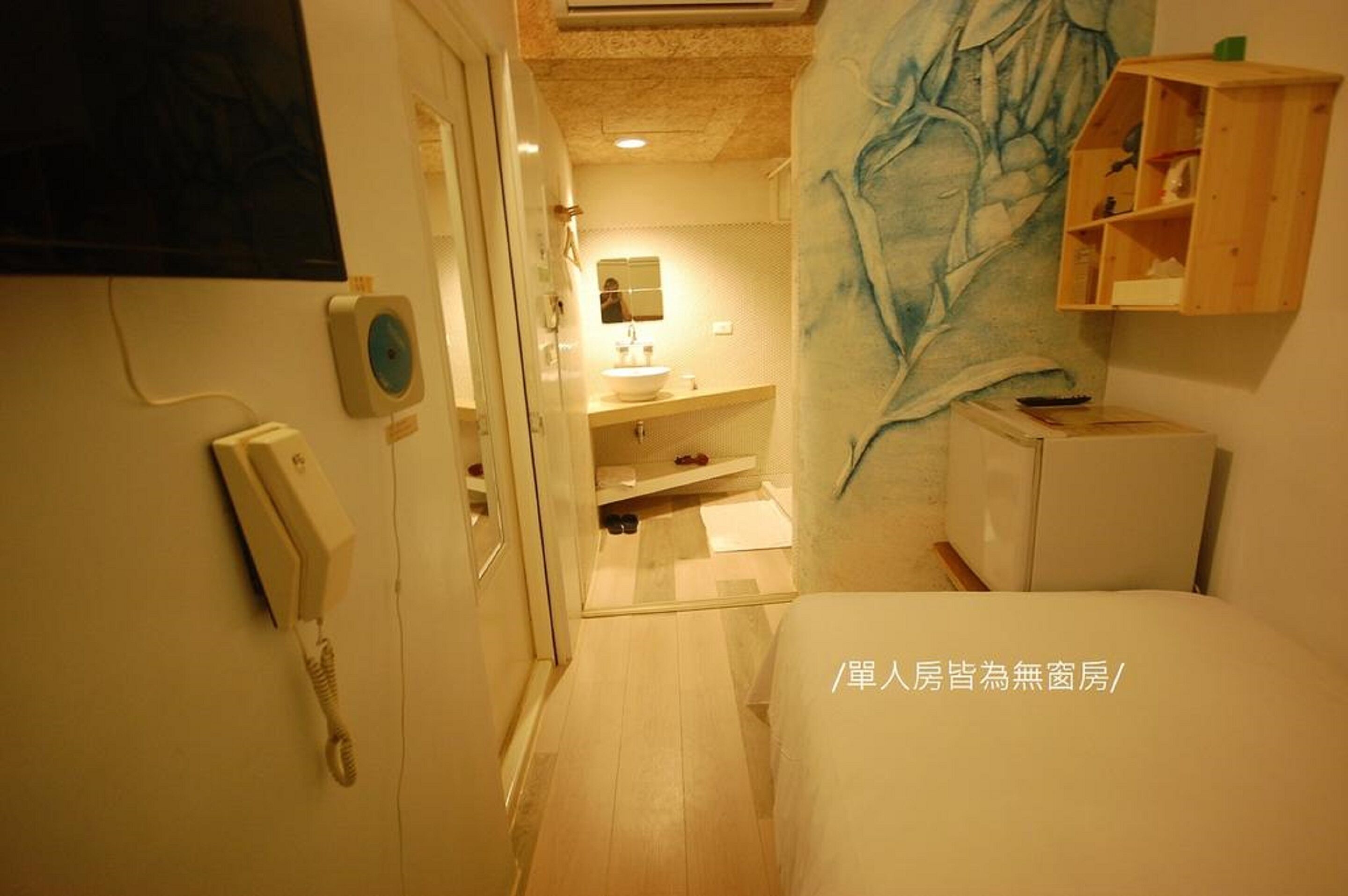 A narrow hotel room with a bed, a small bedside table, a mirror, a wall-mounted shelf, and a telephone.