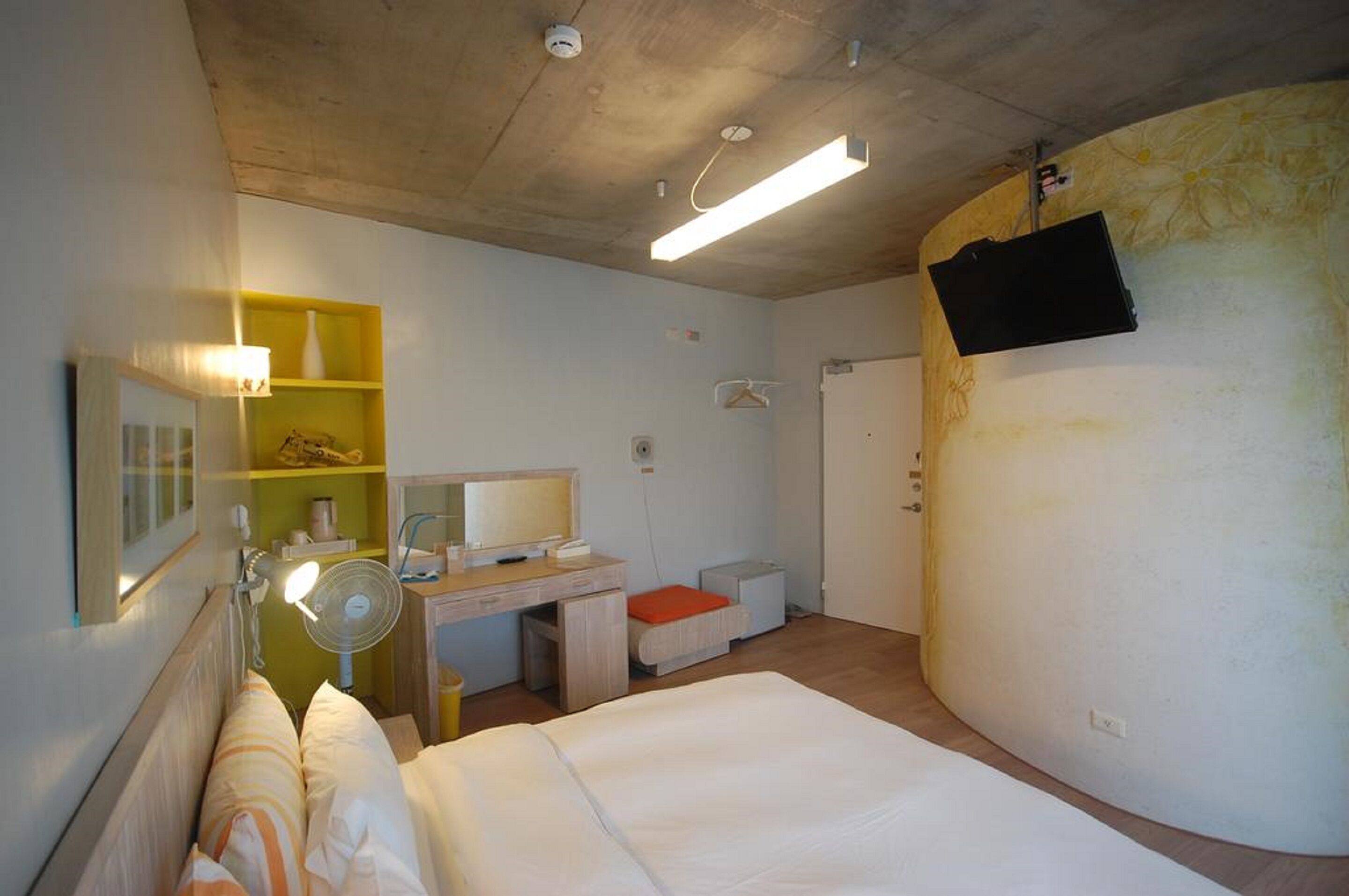 A compact hotel room with a bed, a desk, a chair, a bedside table, a lamp, a mirror, and a TV mounted on the wall.