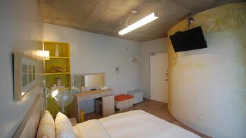 A compact hotel room with a bed, a desk, a chair, a bedside table, a lamp, a mirror, and a TV mounted on the wall.