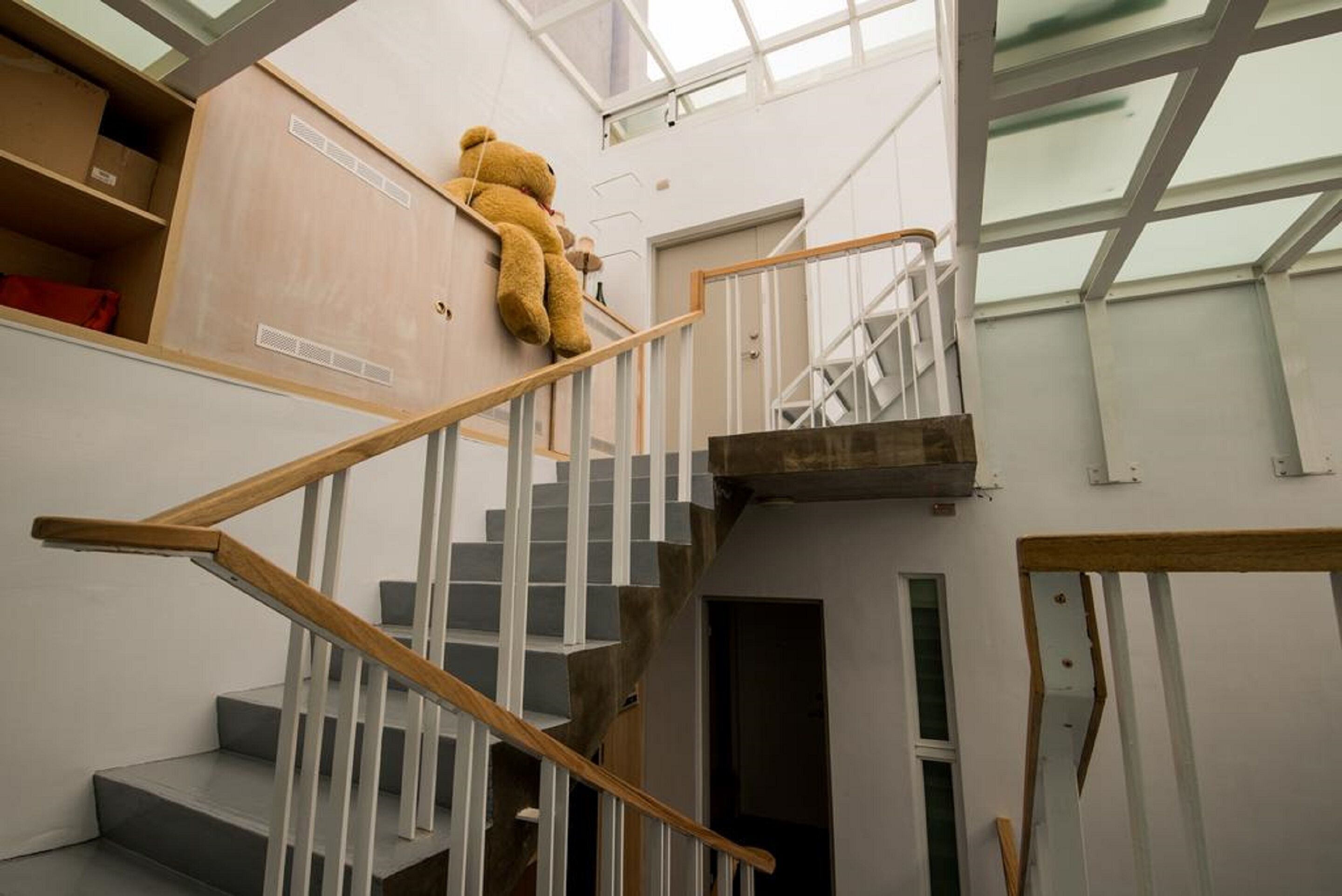 Uma escadaria com corrimão de madeira e balaústres brancos, conduzindo a um patamar onde há um urso de pelúcia grande.