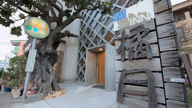 Entrada de edificio con un letrero de madera, un árbol grande y un cartel con caracteres japoneses.