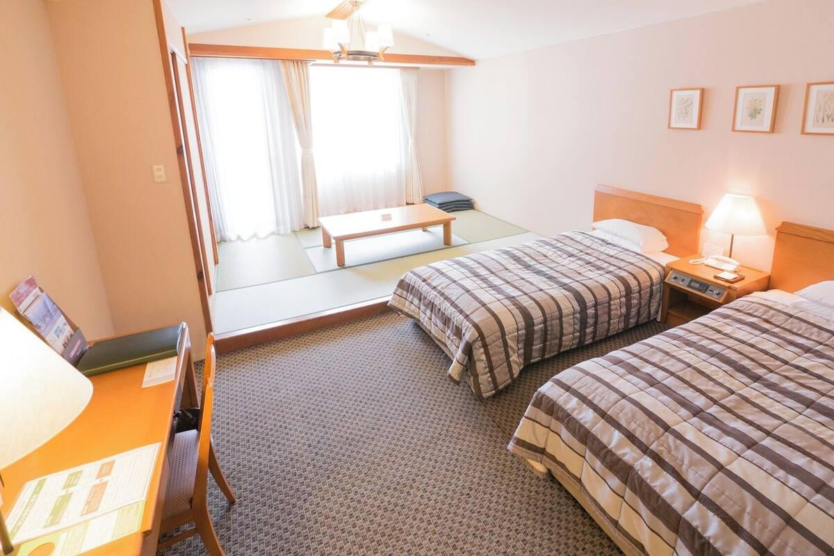 Ein Hotelzimmer mit zwei Betten, einem Schreibtisch mit Lampe, einem Sessel, einem kleinen Tisch und einem Spiegel.