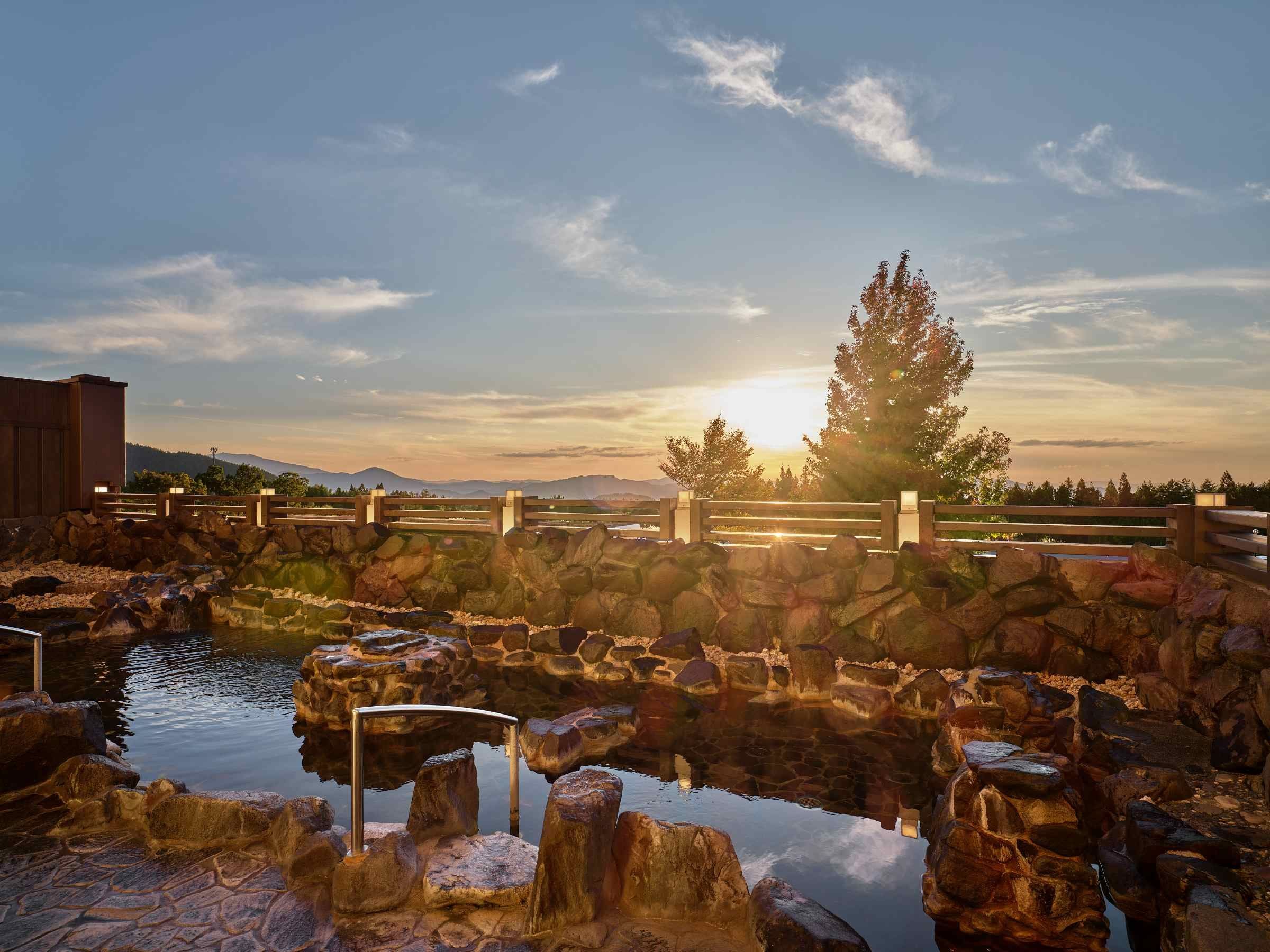 A stone bridge over a reflective water body, a wooden fence, and a tree with the sun setting in the background.