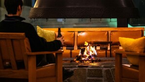 A person sitting by a fireplace with a stone hearth, wooden chairs, and a large black chimney.