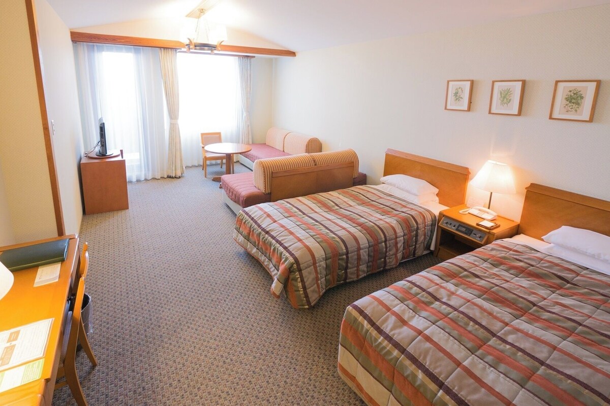A hotel room with two beds, a desk, a chair, a small table, and a window with curtains.