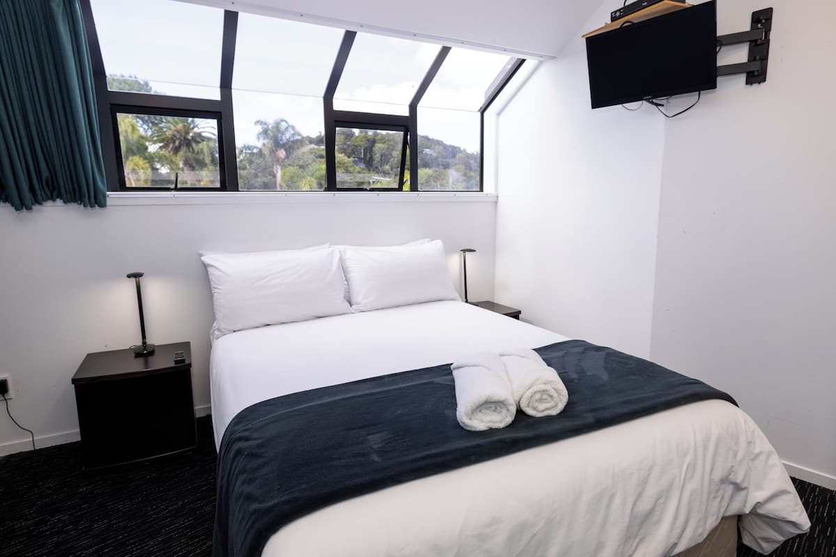 Una habitación de hotel con una cama, televisión montada en la pared, ventana con cortinas y mesita de noche con lámpara.