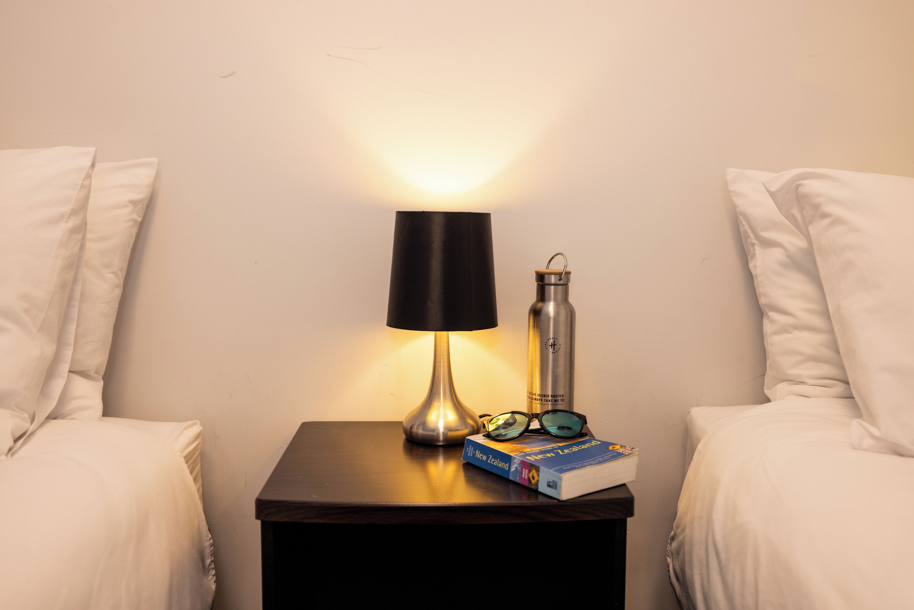 A neatly made bed with white linens, a small bedside table with a lamp, a book, and a bottle.