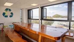Una habitación moderna con una gran mesa de madera, taburetes coloridos y vistas a un cuerpo de agua a través de amplias ventanas.