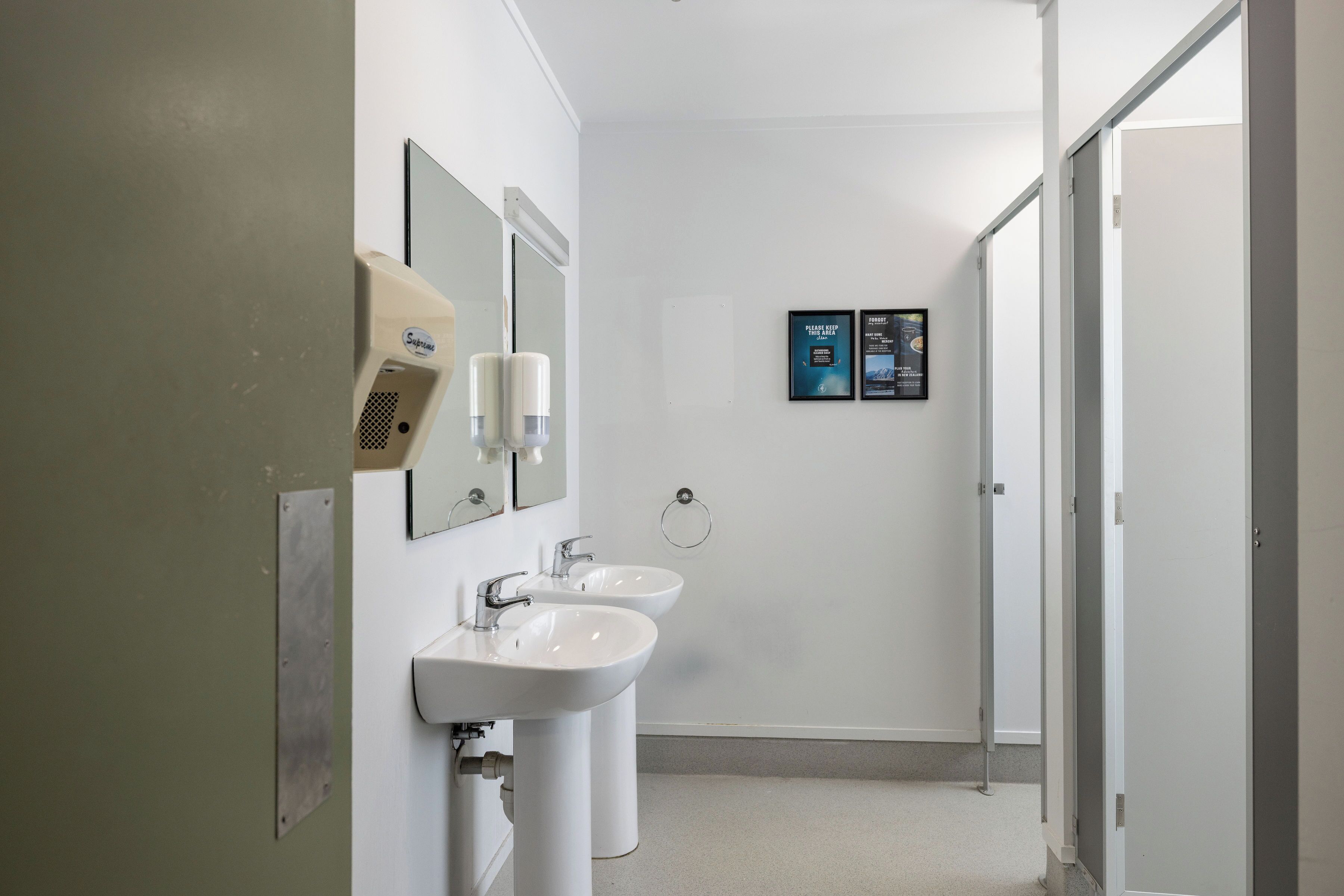 A bathroom with two sinks, a hand dryer, and a mirror. There are two informational posters on the wall.