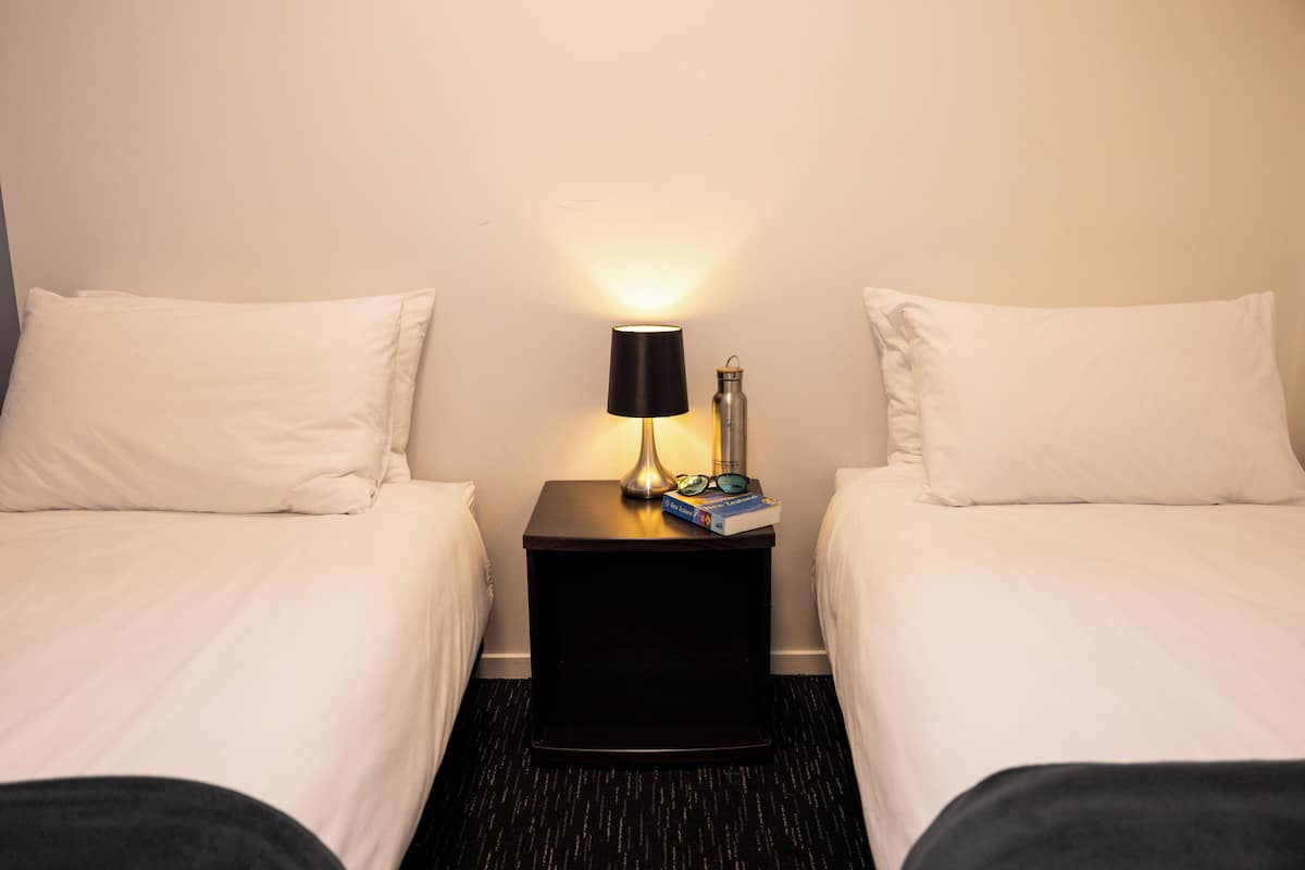 Habitación de hotel con dos camas individuales, una mesita de noche con lámpara, una botella de agua y un par de gafas.