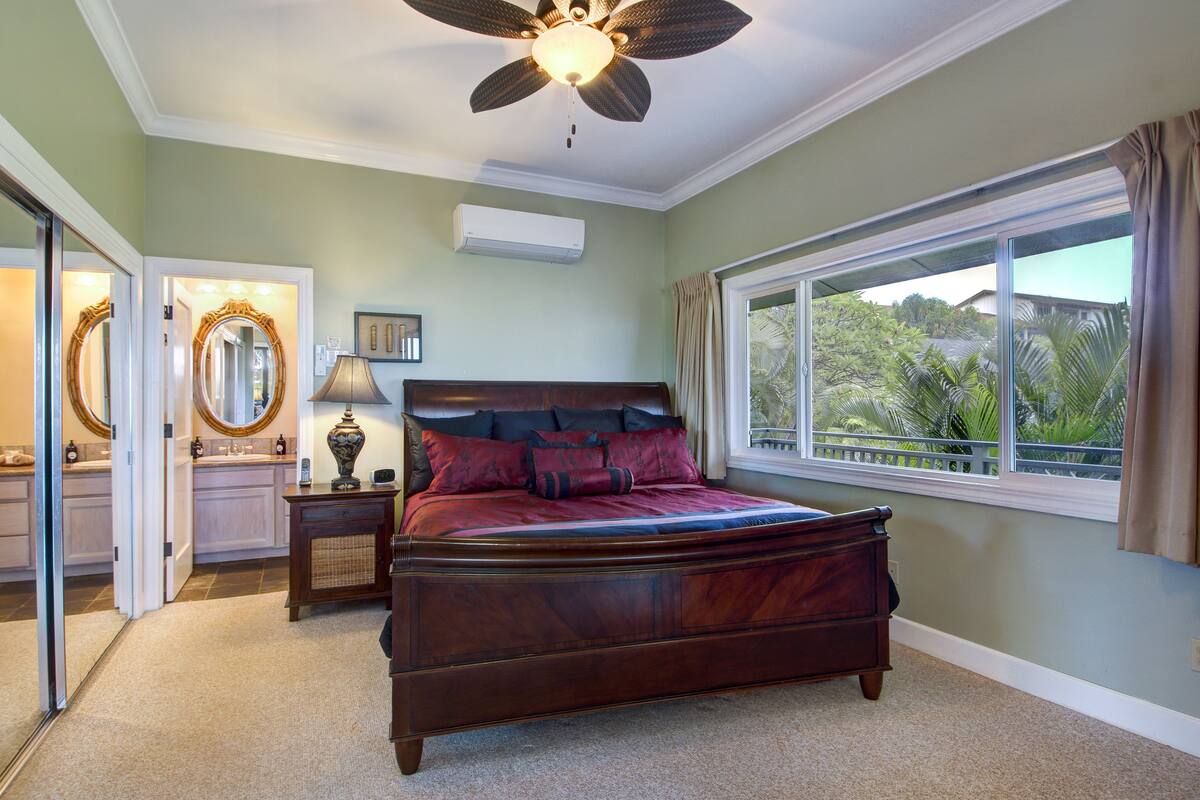 A bedroom with a large bed, a ceiling fan, a window with curtains, and a view of greenery outside.