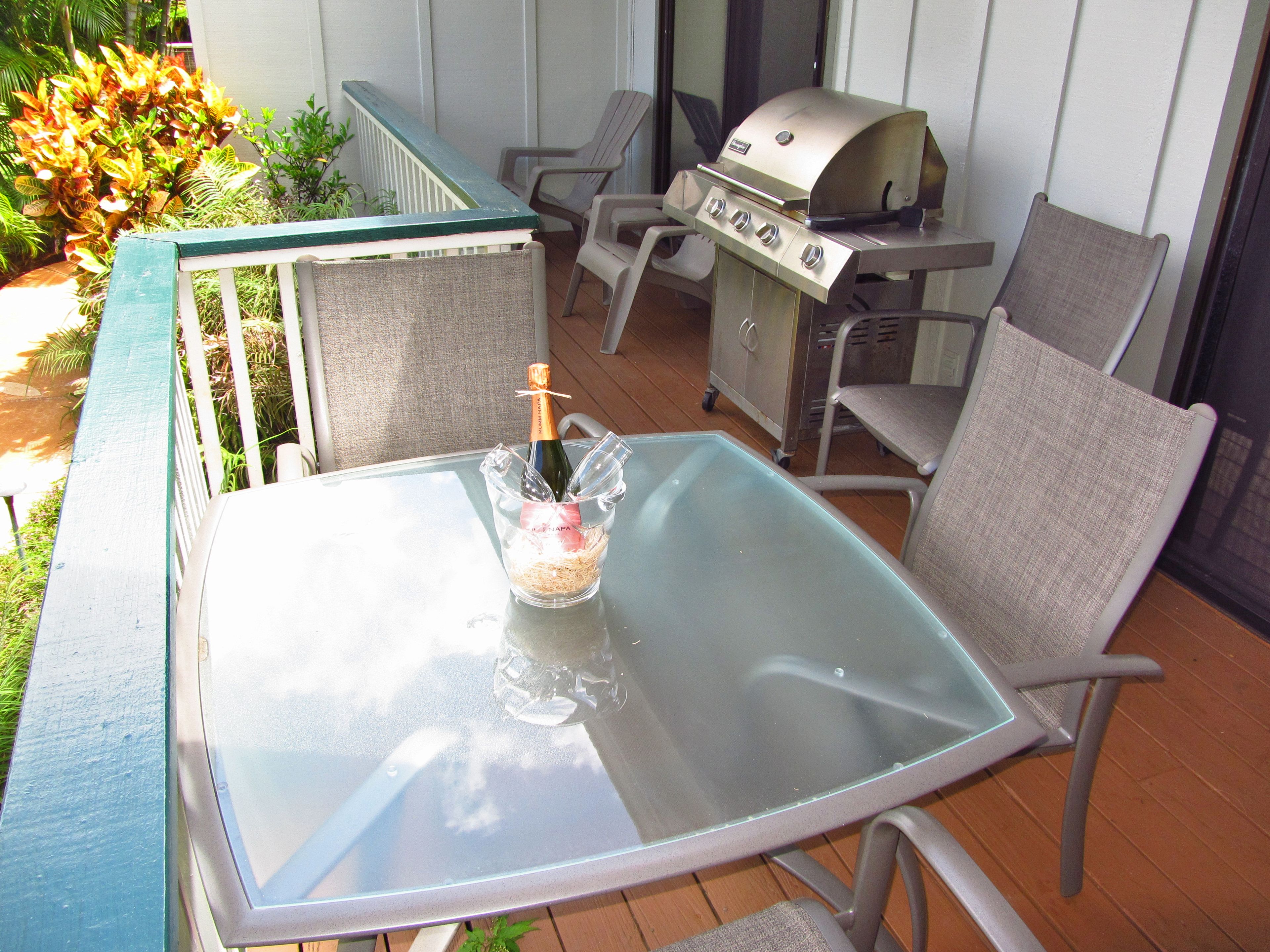 A patio with a glass table, metal chairs, a grill, and a bottle of champagne.