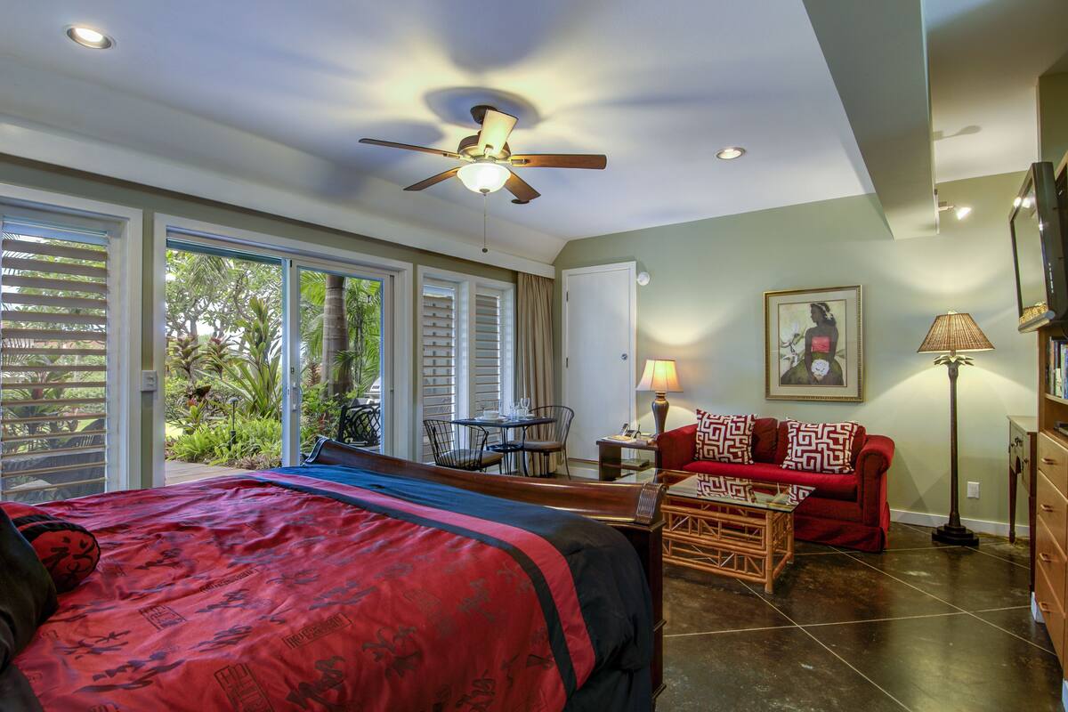 A hotel room with a bed, a red sofa, a dining table, a ceiling fan, and a view of the outdoors.