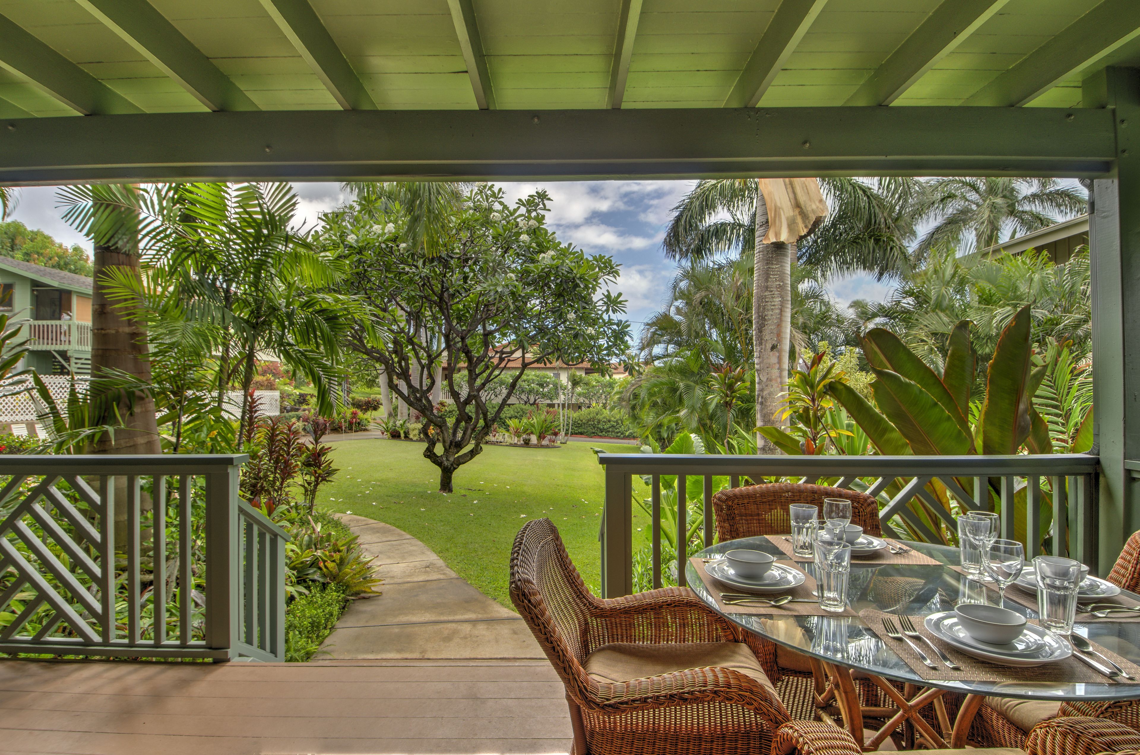 An outdoor dining area with wicker furniture and a glass table, surrounded by lush greenery and a view of a garden.