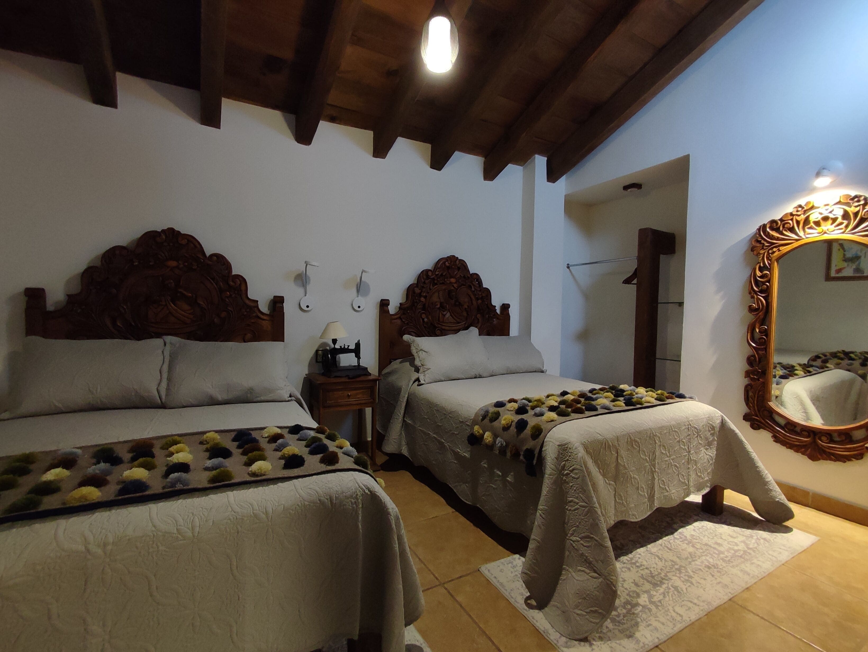 A bedroom with two beds, wooden headboards, a nightstand with a lamp, a mirror, and a door leading to another room.