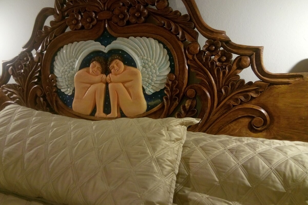 Un lit avec une couette blanche matelassée et une tête de lit en bois ornée de motifs complexes et d’une sculpture représentant un ange.