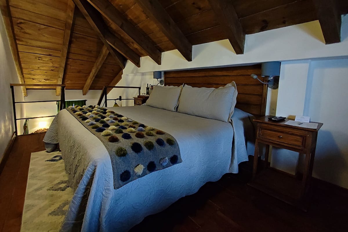Makuuhuone, jossa on puinen katto, kuviollinen peitto sängyllä, yöpöytä lampun kanssa ja pieni sivupöytä.