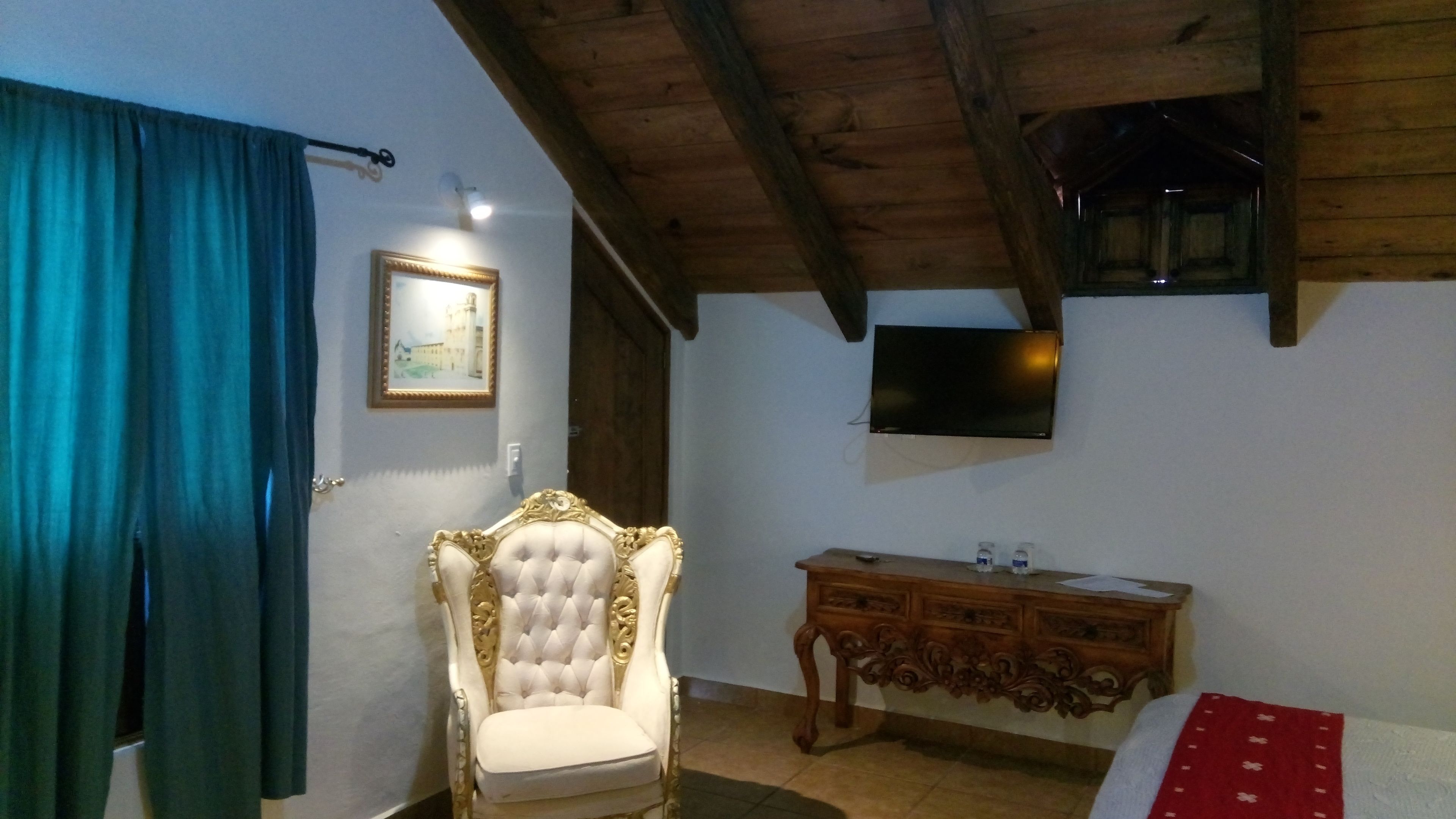 Una habitación con techo de madera, una silla tapizada de blanco, un aparador de madera, un televisor de pantalla plana montado en la pared y una cortina azul.