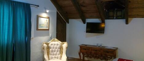 A room with a wooden ceiling, a white upholstered chair, a wooden dresser, a flat-screen TV mounted on the wall, and a blue curtain.