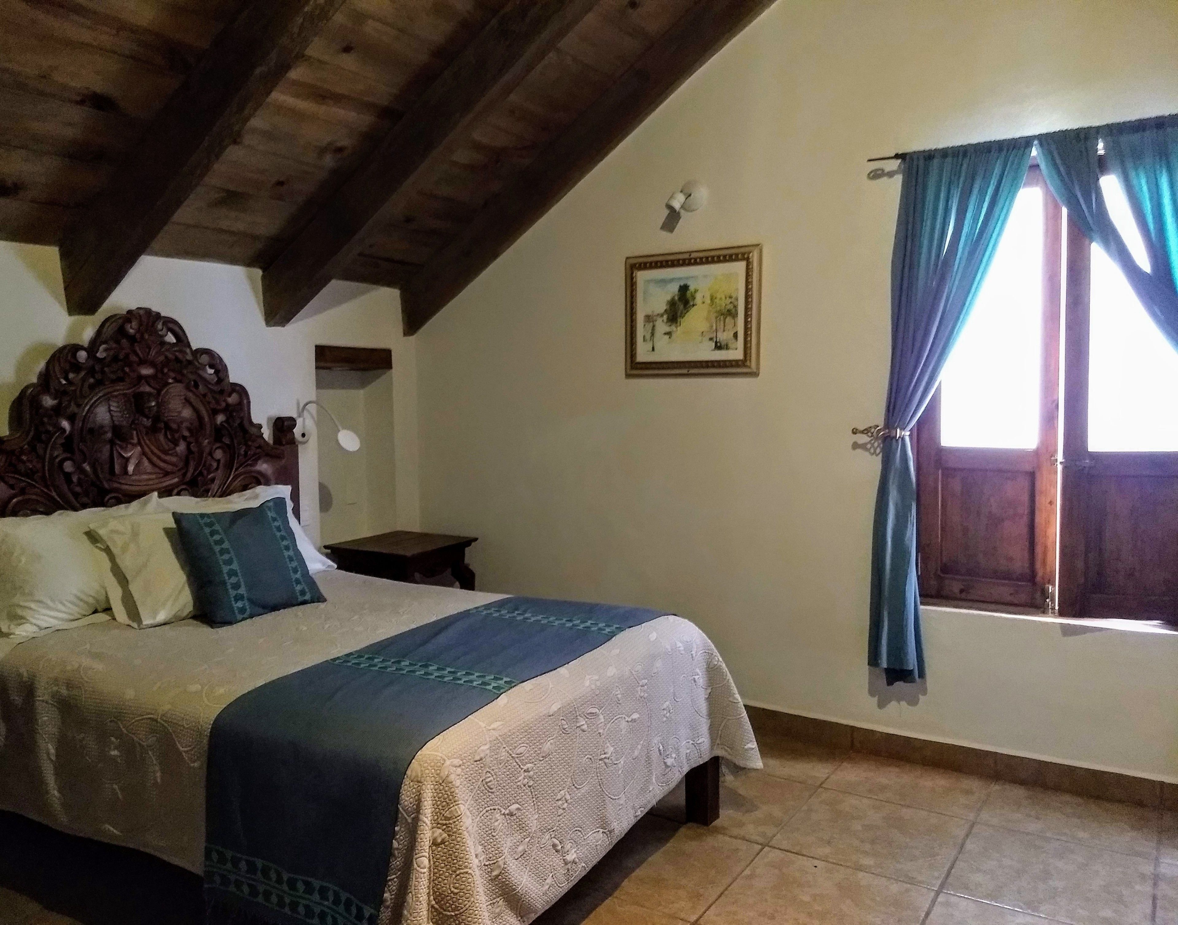 A bedroom with a wooden headboard, a bed with patterned bedding, a small bedside table, a wall-mounted lamp, a framed picture, and a window with curtains.