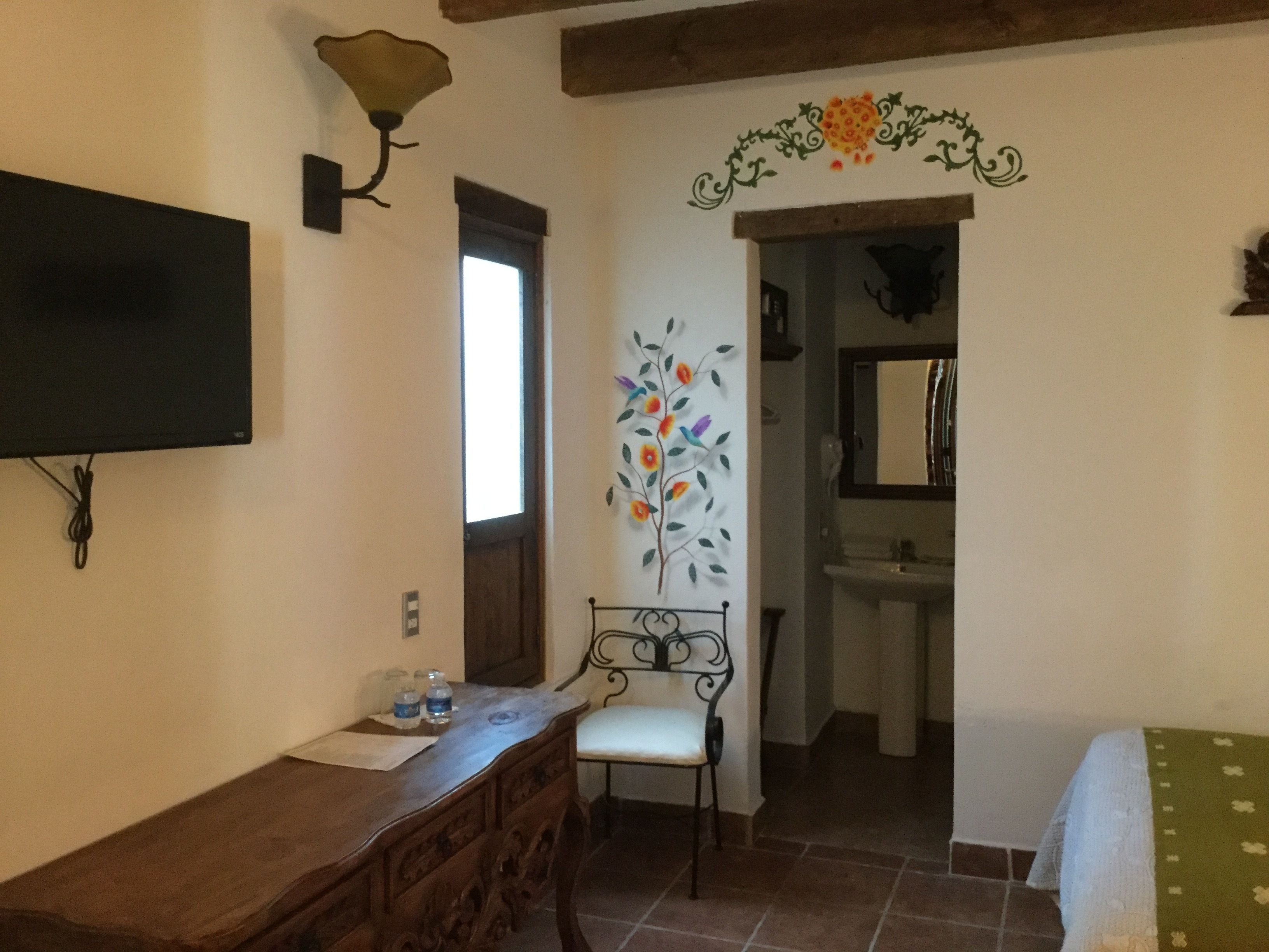 Une pièce comprenant une commode en bois, une chaise, un téléviseur fixé au mur, un élément décoratif mural et une porte donnant sur une salle de bain avec un lavabo et un miroir.
