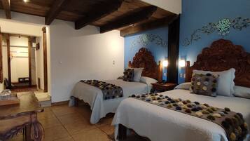 A room with two beds, wooden headboards, a wooden table with a bottle and glasses, and a mirror with floral wall art.