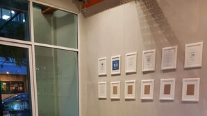 A modern waiting area with a large window, a white desk with an Apple iMac, two purple ottomans, a brown sofa with a blue pillow, and a wall with framed certificates. - SKYPOD Hostel (Kota Kinabalu)