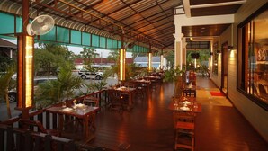 An outdoor restaurant with wooden tables and chairs, a covered roof, and a view of the street. - Boonsiam Hotel (Krabi)