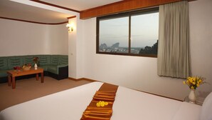 A hotel room with a bed, a green sofa, a small table with a vase of flowers, and a window with a view of the city. - Boonsiam Hotel (Krabi)