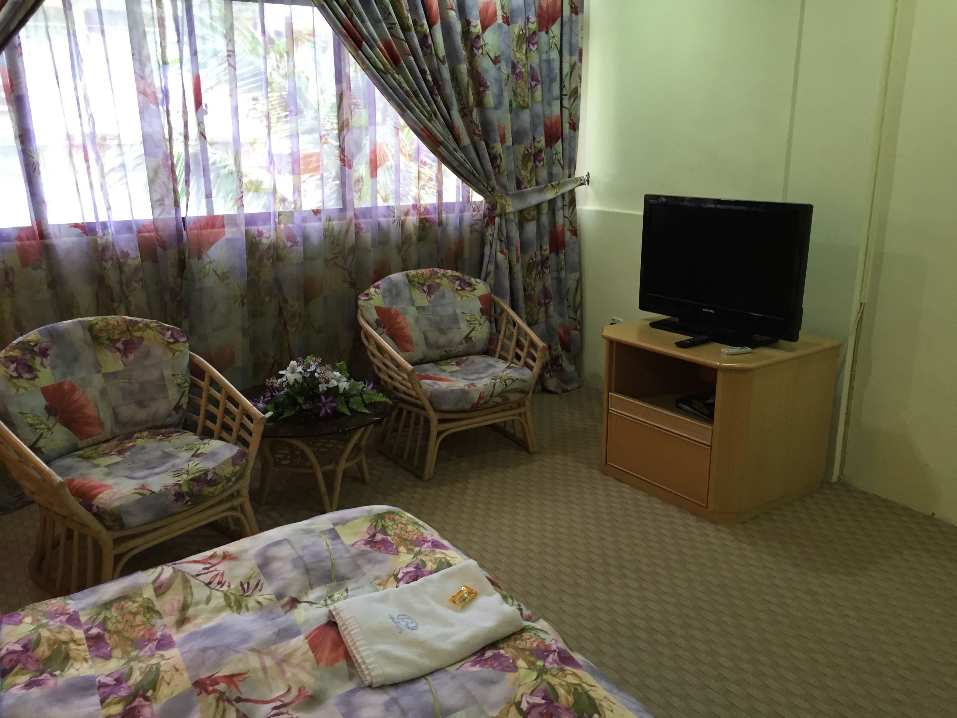Habitación de hotel con una cama, dos sillas, una mesita con flores, un televisor sobre un mueble de madera y cortinas con estampado floral.