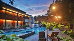 A modern pool area with lounge chairs and a covered seating area, surrounded by a building with warm lighting. - Amata Borobudur Resort (Mungkid)