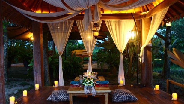 An outdoor dining area with a wooden table set for two, surrounded by candles and draped with white fabric. - Amata Borobudur Resort (Mungkid)