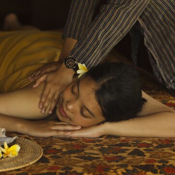 A person receiving a massage on a bed with a patterned bedspread, a small tray with flowers and bottles, and a watch on the masseuse's wrist.