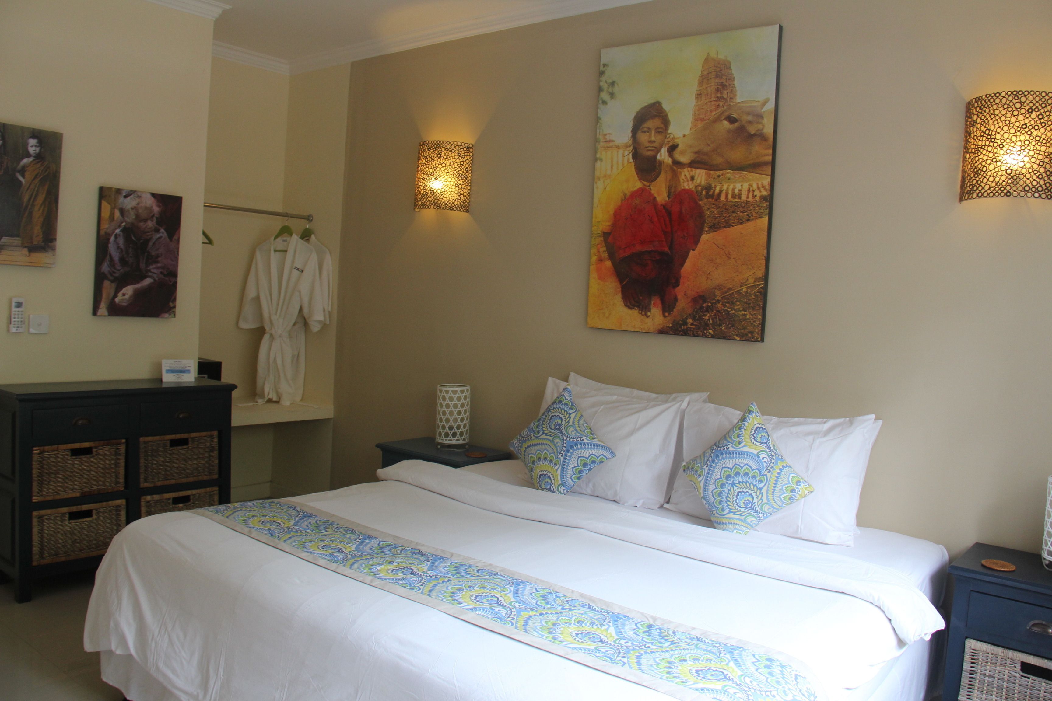 Una habitación de hotel con una cama grande, una mesita de noche, una lámpara, una obra de arte colgada en la pared, un perchero y una pequeña mesa con una lámpara.