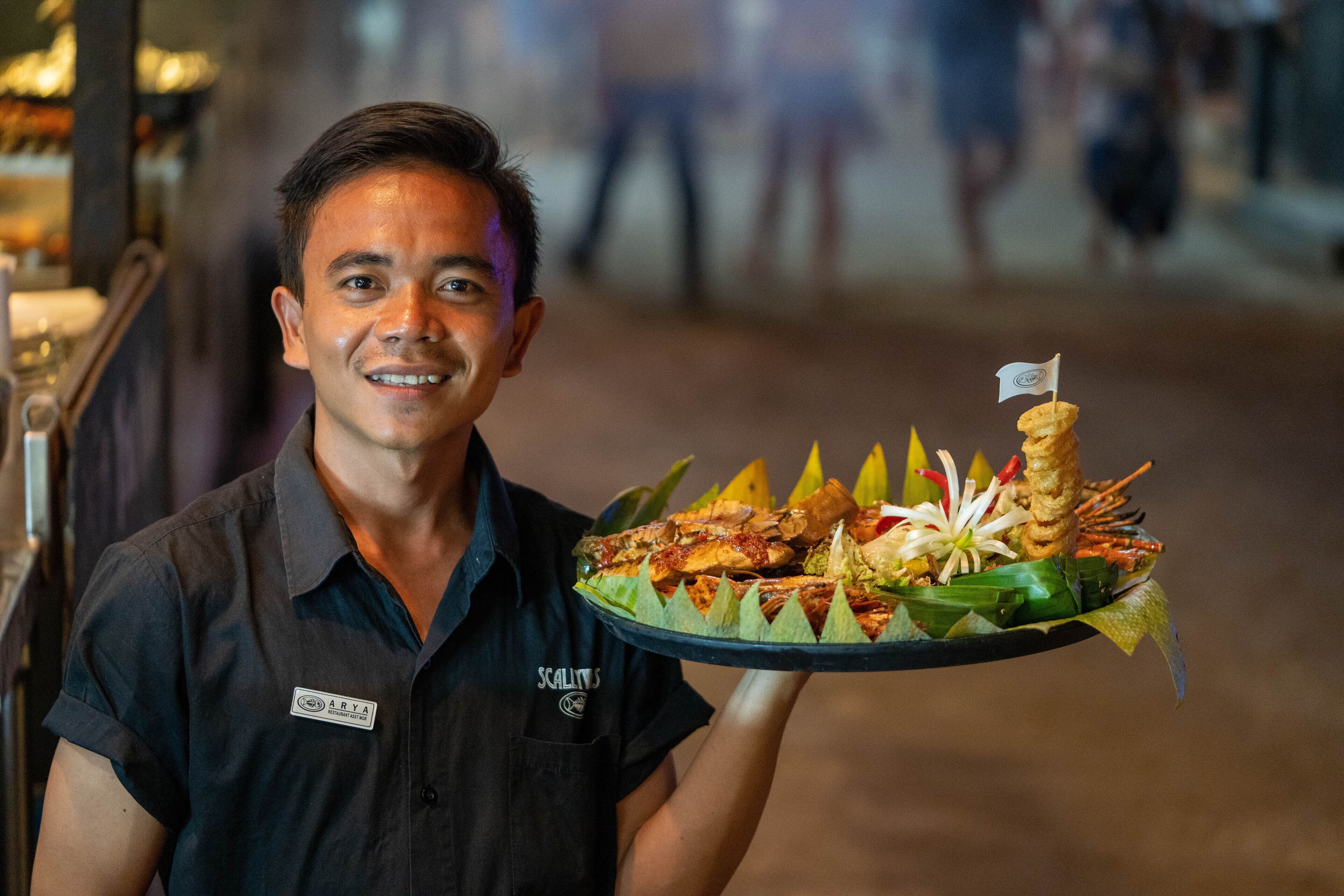Een persoon draagt een dienblad met diverse soorten eten, waaronder zeevruchten en groenten, in een omgeving die lijkt op een restaurant of een ruimte voor horeca.