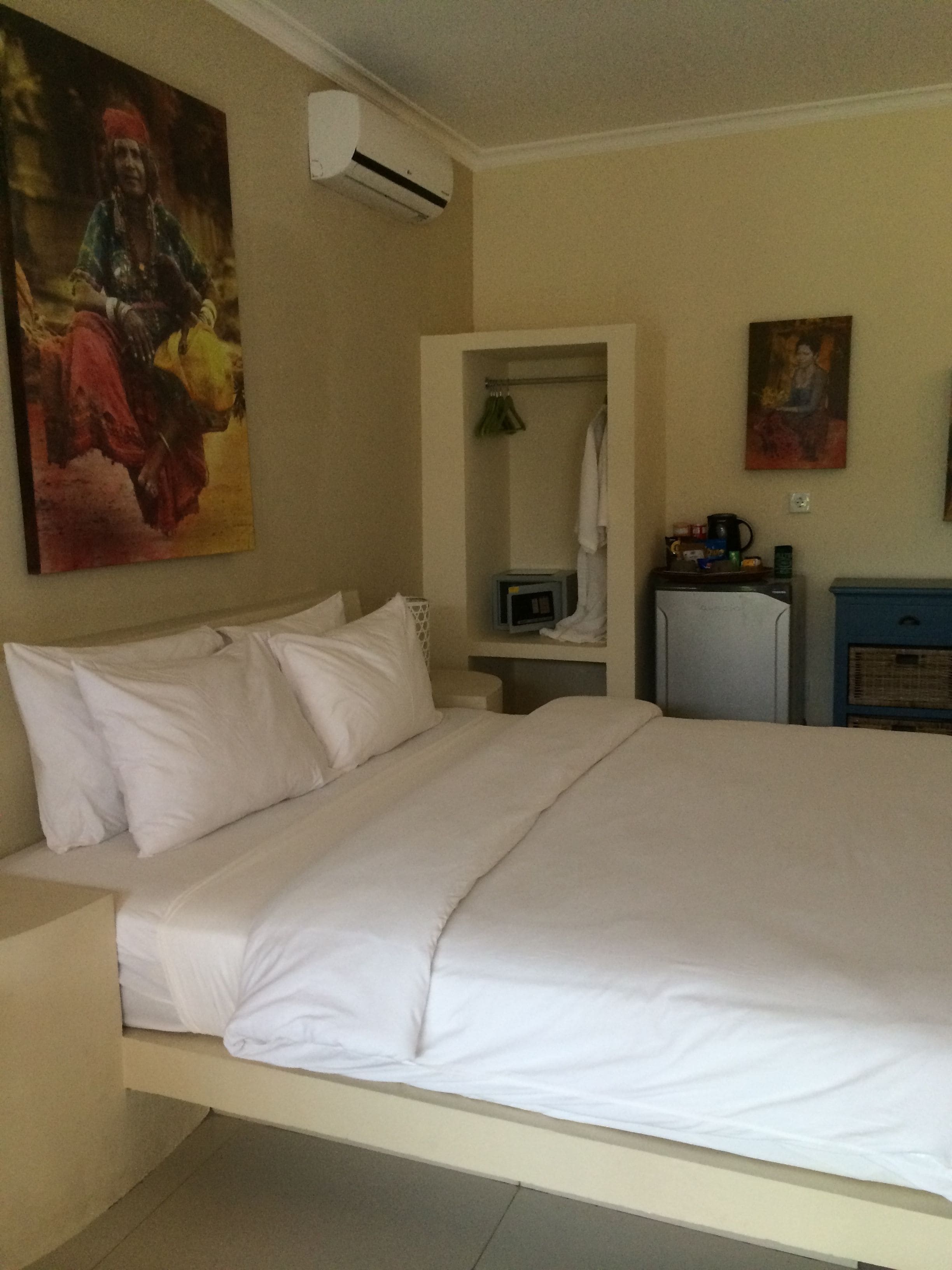 A hotel room with a large bed, a small desk with a computer, a microwave, and a refrigerator. There is a painting on the wall and an air conditioning unit above the bed.