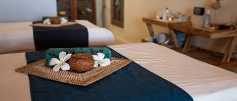 Une chambre d’hôtel avec deux lits, chacun orné d’un oreiller bleu foncé et d’un plateau en osier décoré de fleurs. On aperçoit en arrière-plan une armoire en bois et une petite table sur laquelle sont posées des bougies.