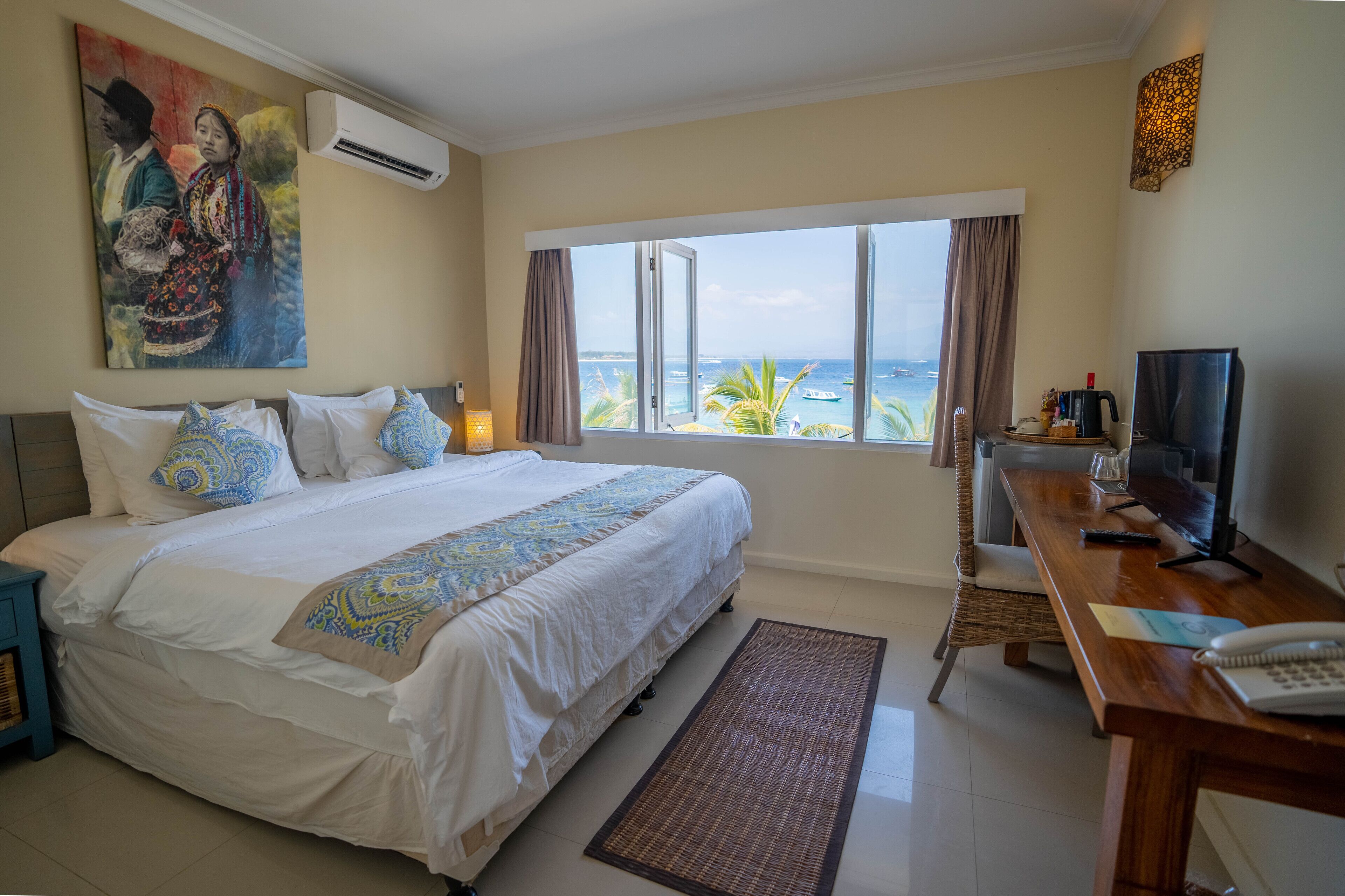 A hotel room with a large bed, a desk with a computer, a window with a view of the sea, and a painting on the wall.