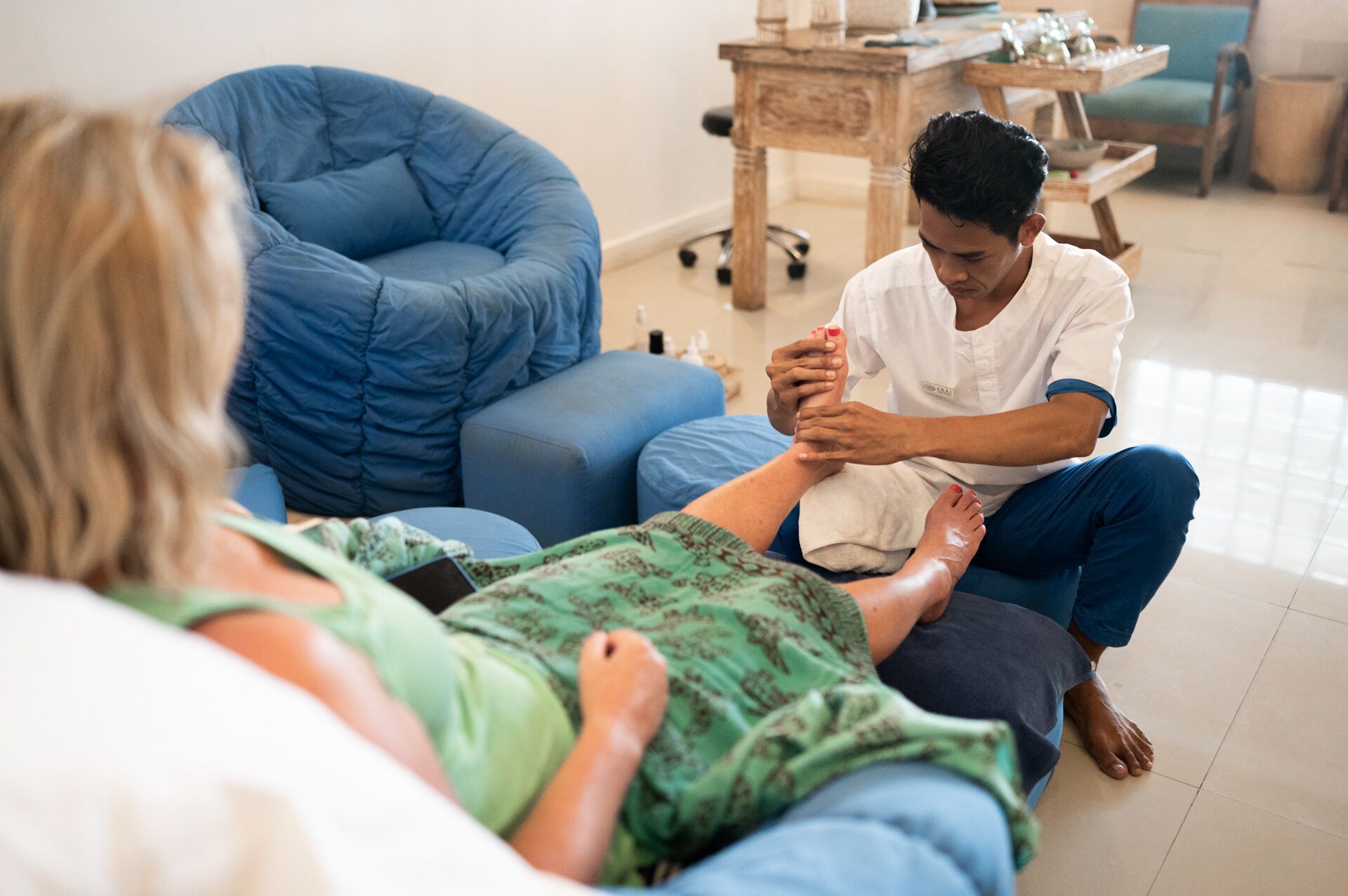 A person receiving a foot massage in a room with a blue bean bag chair and a wooden table.