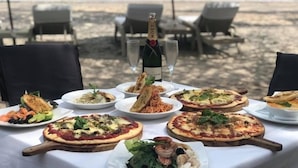 A table set on a beach with various dishes, including pizzas and seafood, and a bottle of champagne. - Scallywags Resort (Gili Trawangan)
