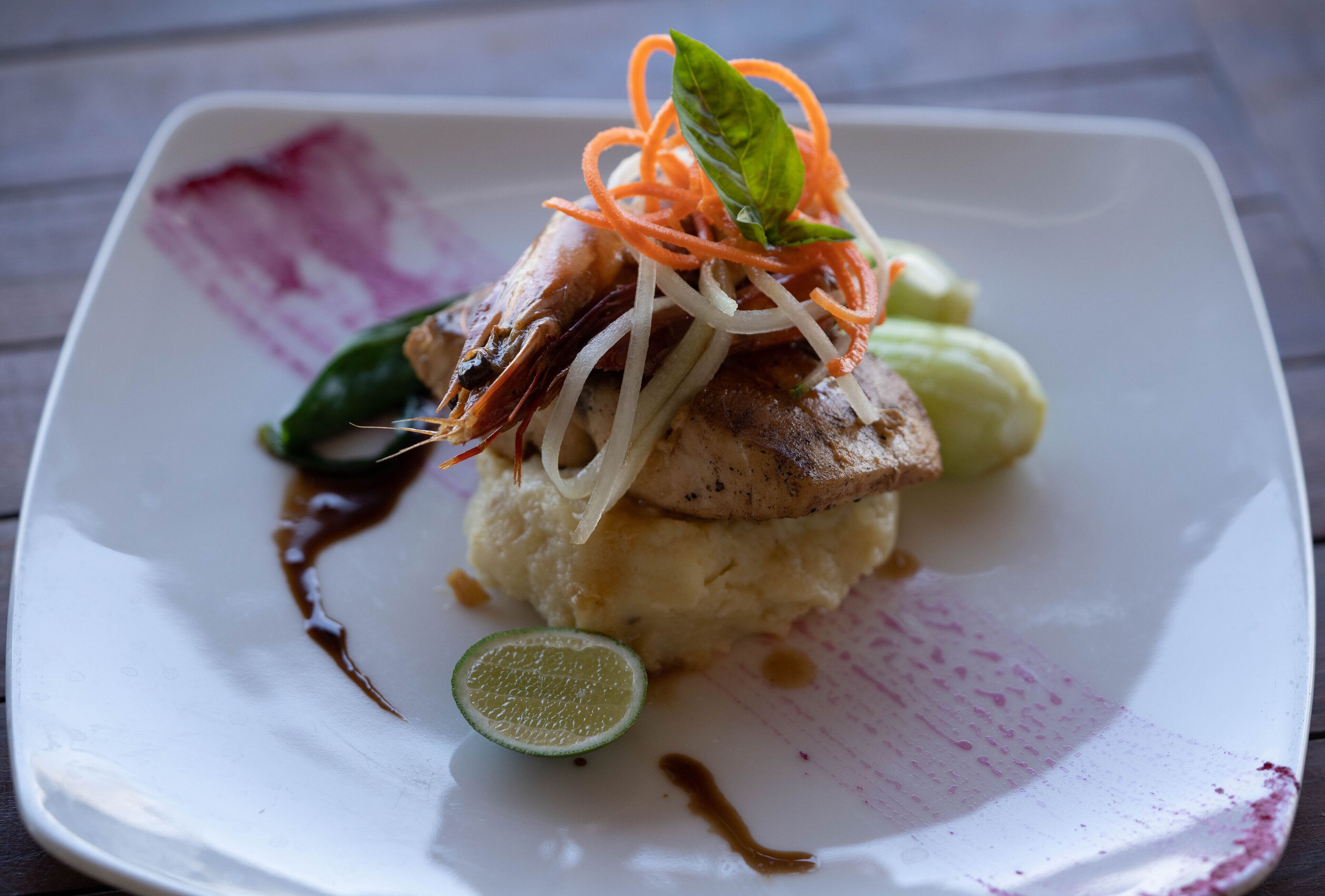 Une assiette carrée contenant un plat composé d’une viande grillée, possiblement du poulet, servie sur une purée de pommes de terre, et agrémentée de juliennes de carottes et d’une feuille de basilic.