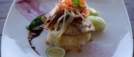 A square plate with a dish consisting of a grilled item, possibly chicken, atop mashed potatoes, garnished with carrot strips and a basil leaf.