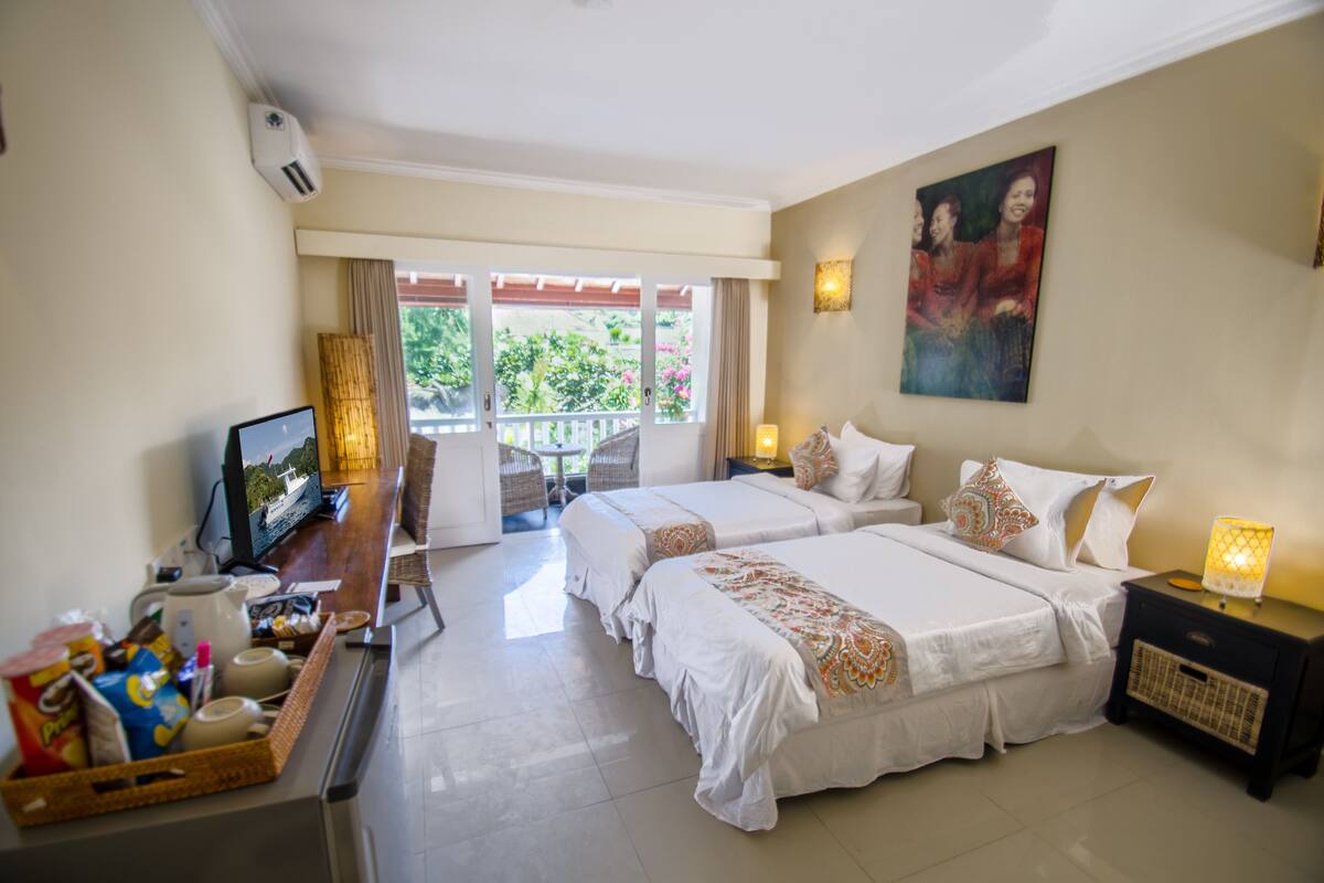 Une chambre d’hôtel avec deux lits, un bureau avec un ordinateur, une télévision et un balcon donnant sur un espace verdoyant.