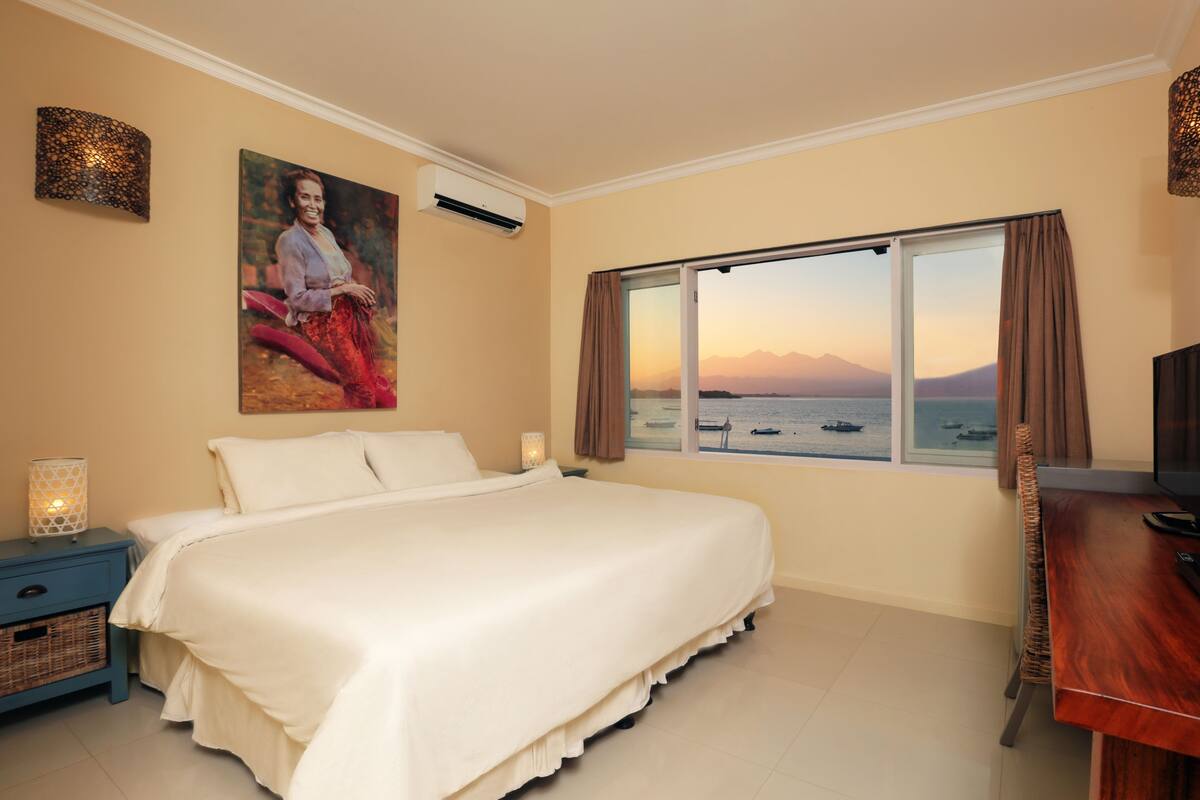 Une chambre avec un grand lit, une table de chevet avec une lampe, un bureau avec une chaise, un tableau au mur et une fenêtre donnant sur la mer et les montagnes.