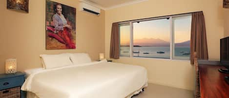 Un dormitorio con una cama grande, una mesita de noche con lámpara, un escritorio con silla, un cuadro en la pared y una ventana con vistas al mar y a las montañas.