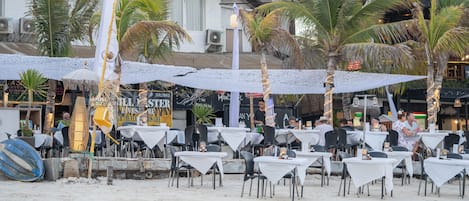 Un espacio de comedor al aire libre con mesas y sillas, palmeras y un edificio al fondo.